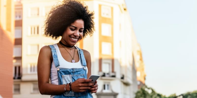 A woman walking on the street looks down at her mobile phone and smiles.