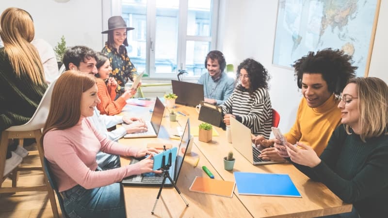 A group of people sit at a table, smiling, as they brainstorm and work on their laptops.